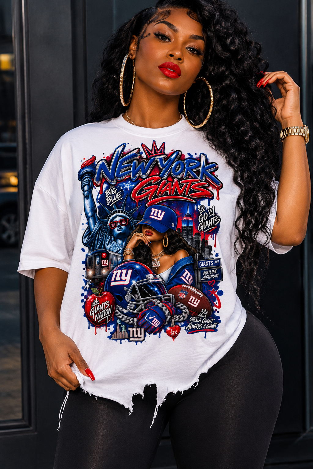 A woman wearing a white graphic t-shirt featuring the New York Giants logo and sports-themed artwork, paired with black leggings, standing in front of a dark doorway.