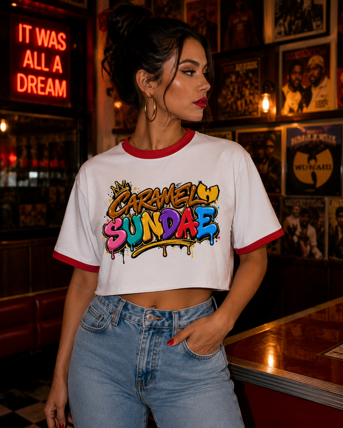 A woman wearing a white ringer crop top t-shirt with red trim and a colorful graffiti-style graphic that reads 'Garmely Sundae'. She is posing in a dimly lit, warm-toned retro bar with vinyl records on the wall.