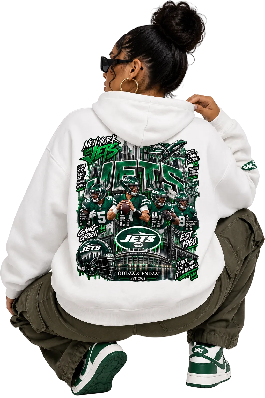 A person wearing a white hoodie with a large green and black New York Jets graphic on the back, crouching in olive cargo pants and green-and-white sneakers against a light gray studio background.