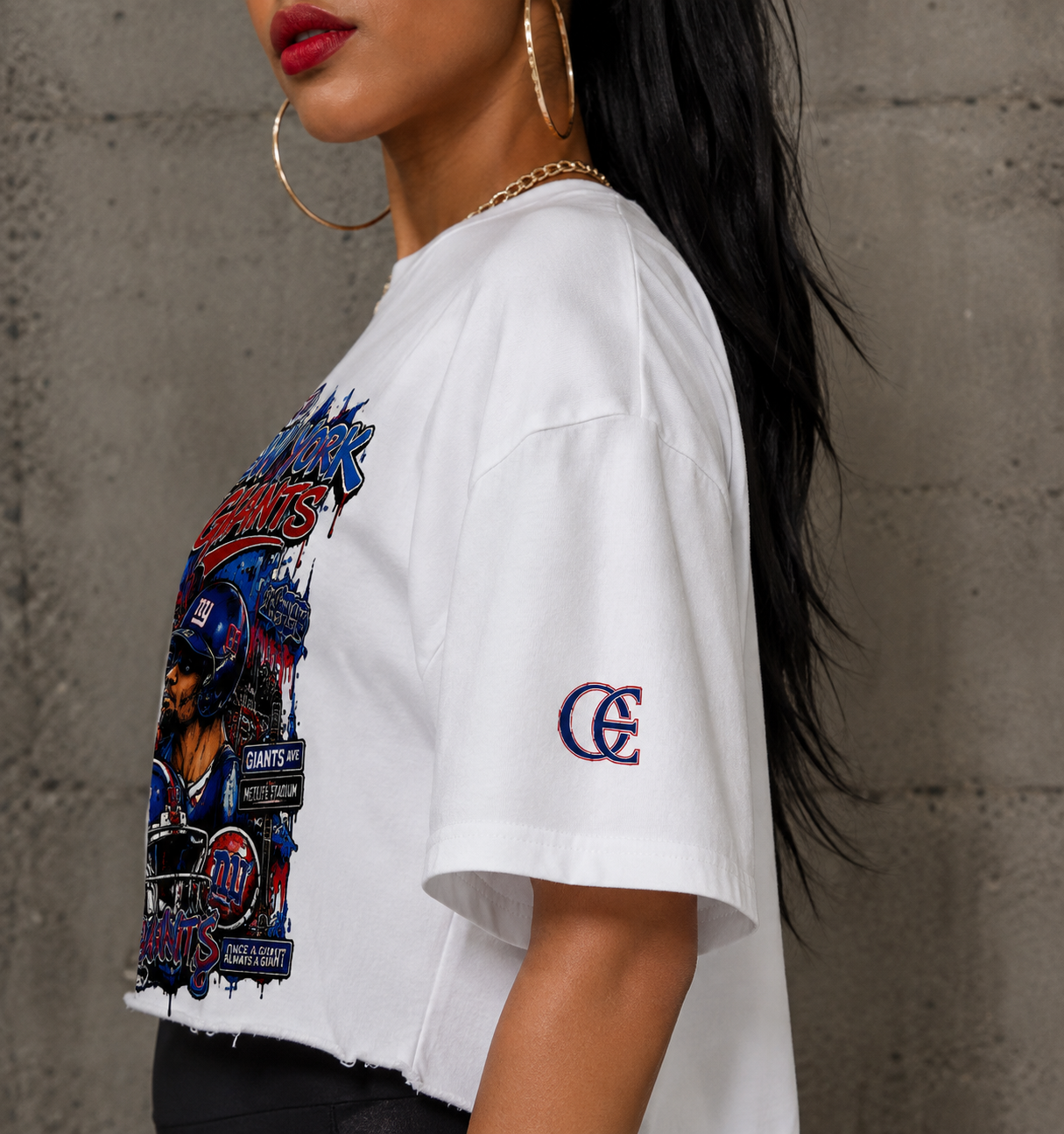 A woman wearing a white cropped t-shirt with a large blue and red New York Giants graphic on the front and a blue embroidered 'GE' logo on the sleeve, posed against a gray concrete wall.
